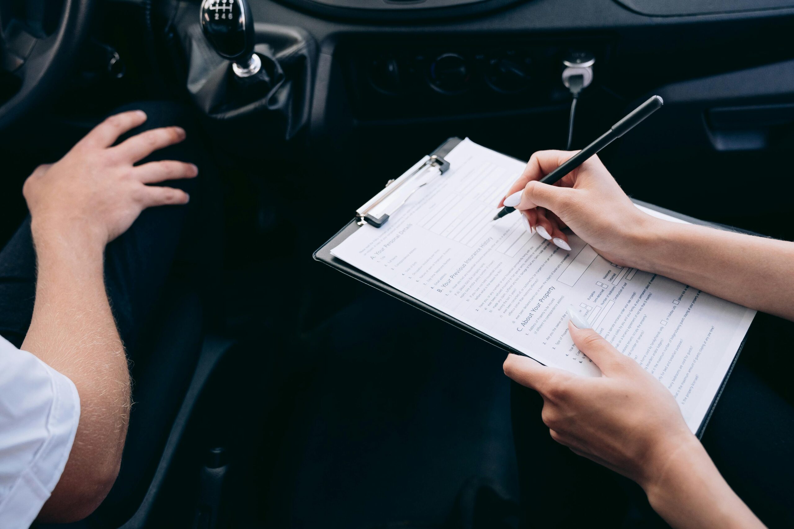 Home Close-up of a person writing on a clipboard inside a car, showing hands and a gear shift.